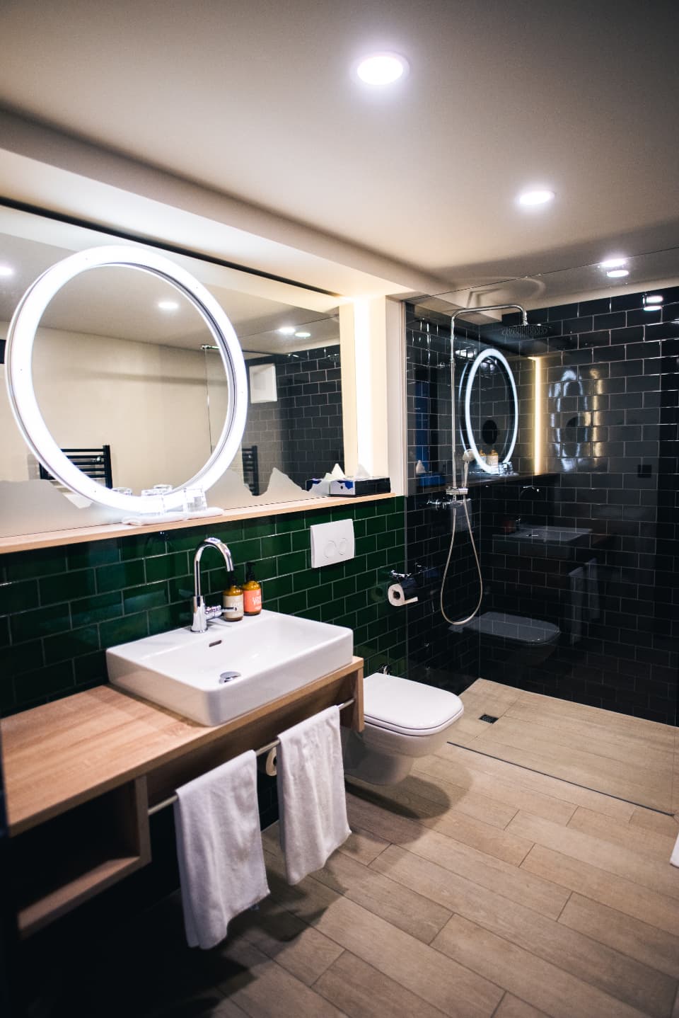 A dark-tiled hotel bathroom with a round mirror and a basin