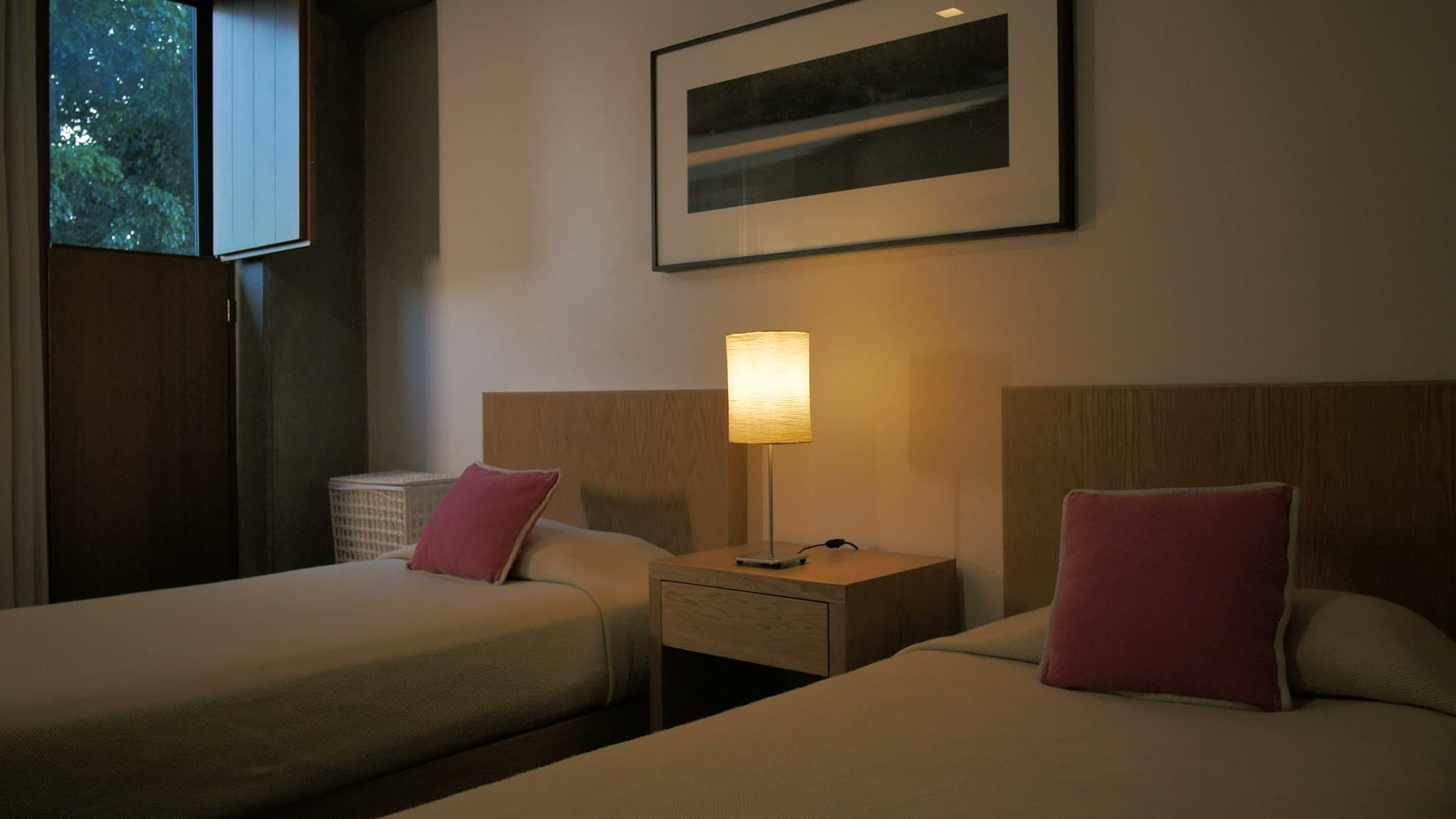 A twin room at dusk: two made beds, a lit lamp between them and a window onto trees