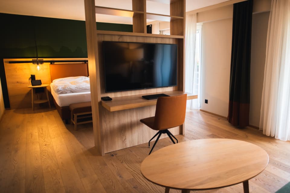 A hotel studio with a desk, a wall-mounted screen and a made bed by the window