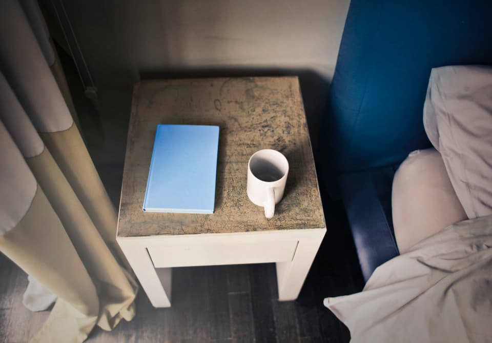 A bedside table with a book and a cup beside a bed with blue walls